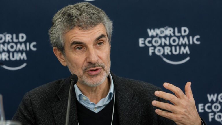 Hombre hablando frente a un micrófono, mano derecha levantada; fondo azul con el logotipo 'World Economic Forum'.