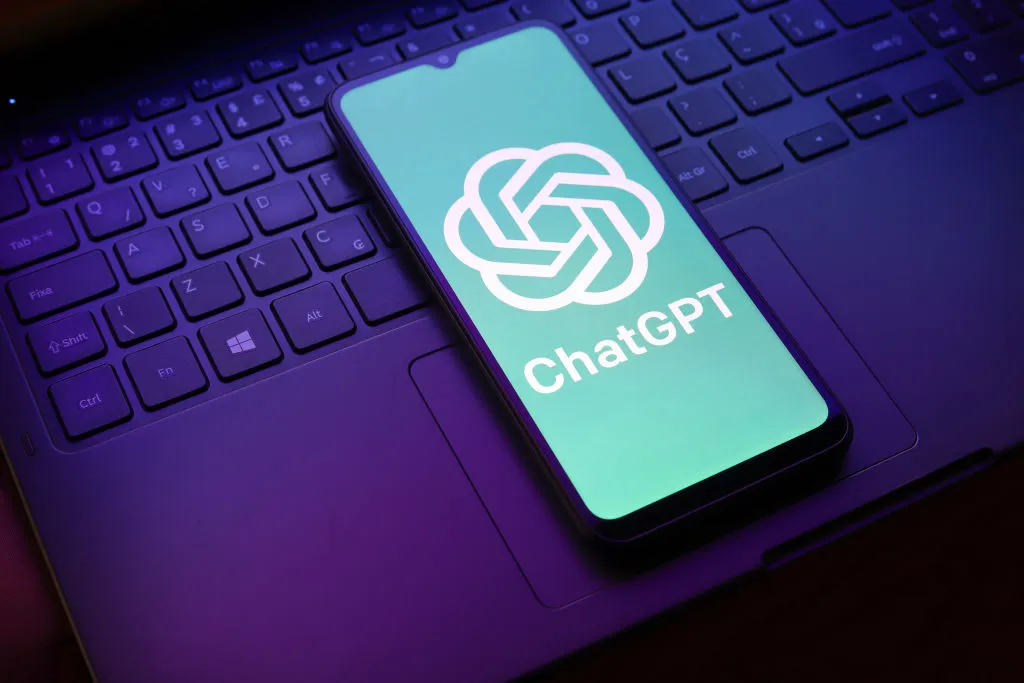 Teléfono con el logotipo y el nombre de ChatGPT en pantalla, colocado sobre el teclado de un portátil con iluminación violeta.