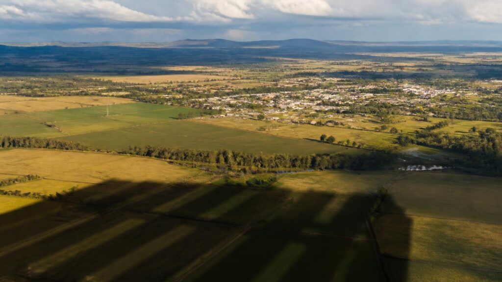 Vista aérea de campos agrícolas y una localidad al fondo; sombras rectangulares alargadas cruzan las parcelas bajo un cielo parcialmente nuboso.