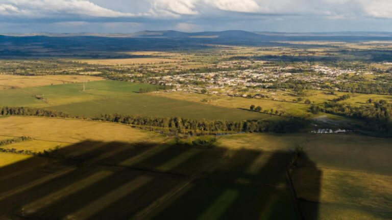 Vista aérea de campos agrícolas y una localidad al fondo; sombras rectangulares alargadas cruzan las parcelas bajo un cielo parcialmente nuboso.