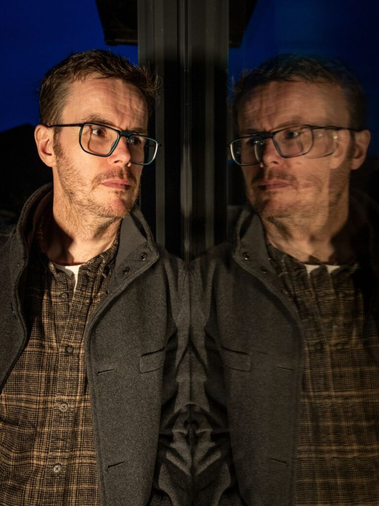 Benjamin Skuse, con gafas, junto a una ventana que refleja su rostro de noche.